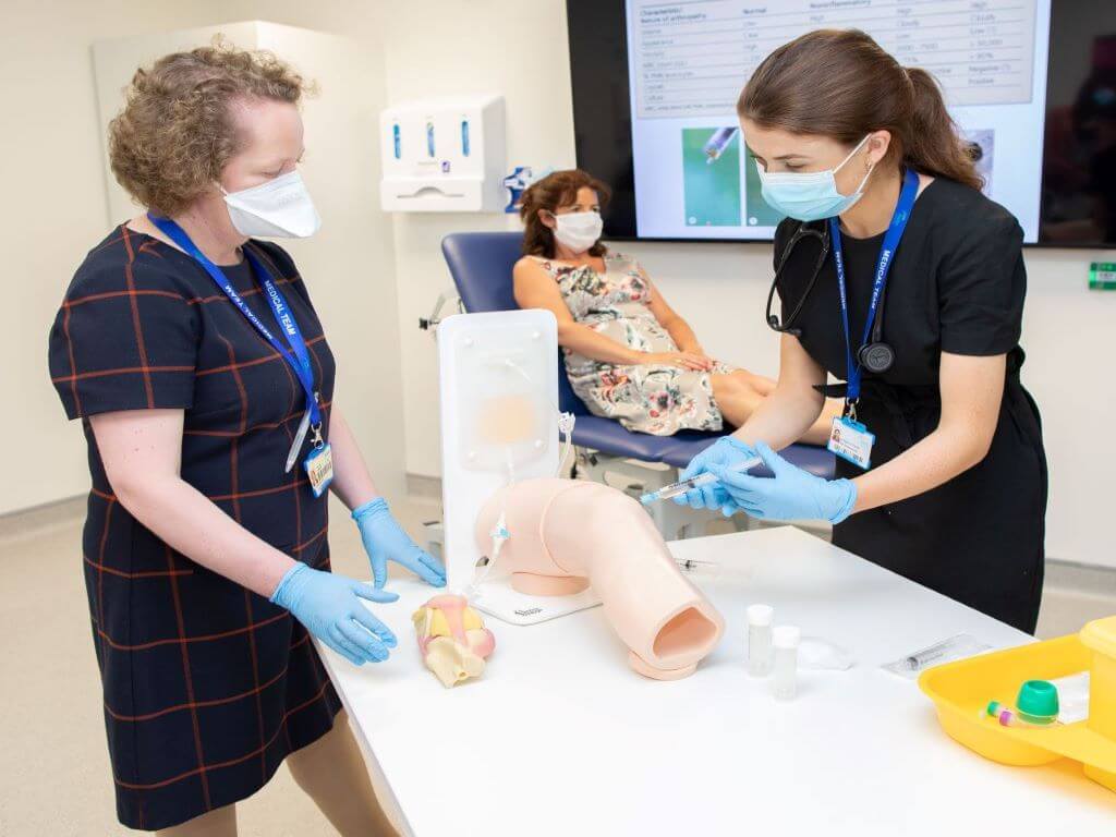 Dr Bernadette Lynch, Consultant Rheumatologist at University Hospital Galway, guiding a Medical Trainee through a simulated knee aspiration. Patient on stretcher in background. All wearing medical masks.